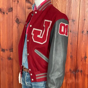 Op de afbeelding: Bordeauxrode varsity jas met grijze leren mouwen. De jas heeft een grote witte "J" op de borst en het nummer "01" op de mouw. Grijze en bordeauxrode gestreepte biezen accentueren de kraag, manchetten en taille.