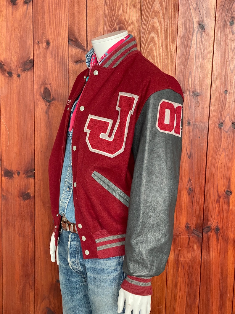 Op de afbeelding: Bordeauxrode varsity jas met grijze leren mouwen. De jas heeft een grote witte "J" op de borst en het nummer "01" op de mouw. Grijze en bordeauxrode gestreepte biezen accentueren de kraag, manchetten en taille.