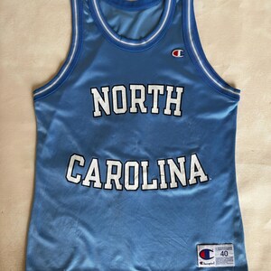 May include: A light blue sleeveless basketball jersey with white and blue striped trim on the collar and armholes. The text NORTH CAROLINA is printed in white with black outlines across the chest. A Champion logo and size 40 tag are visible.