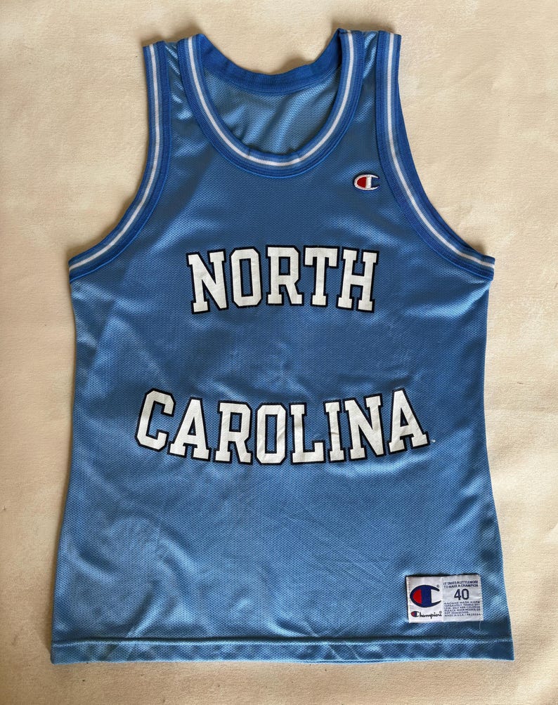 May include: A light blue sleeveless basketball jersey with white and blue striped trim on the collar and armholes. The text NORTH CAROLINA is printed in white with black outlines across the chest. A Champion logo and size 40 tag are visible.