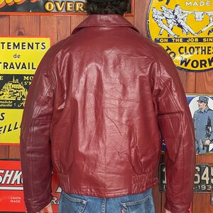 May include: A burgundy leather bomber jacket with a ribbed hem and cuffs. The jacket has a classic collar and a front zipper. The person is wearing a black bowler hat and blue jeans. Vintage clothing.