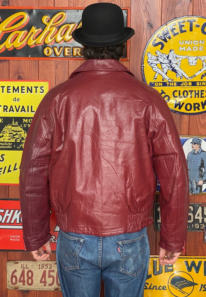 May include: A burgundy leather bomber jacket with a ribbed hem and cuffs. The jacket has a classic collar and a front zipper. The person is wearing a black bowler hat and blue jeans. Vintage clothing.