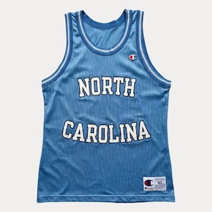 May include: A light blue sleeveless basketball jersey featuring the text NORTH CAROLINA in white block letters with navy blue outlines. The garment includes a white and navy blue striped trim on the neckline and armholes, with a Champion logo patch.