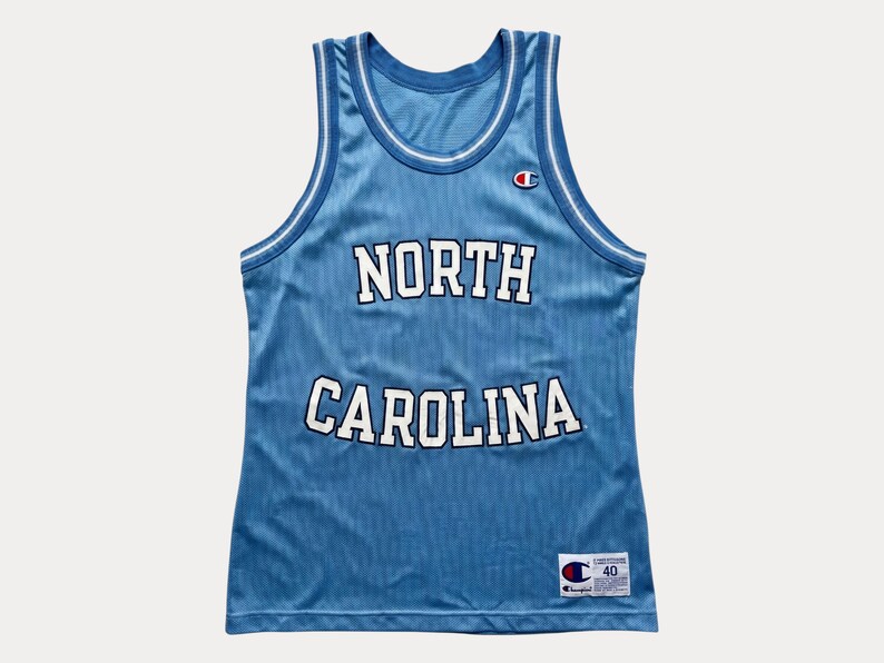 May include: A light blue sleeveless basketball jersey featuring the text NORTH CAROLINA in white block letters with navy blue outlines. The garment includes a white and navy blue striped trim on the neckline and armholes, with a Champion logo patch.