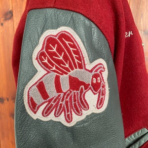 Op de afbeelding: Bordeauxrode varsity jas met een grijze leren mouw. De jas heeft een groot geborduurd embleem van een gestileerde rood-grijze bij. De jas is gemaakt van een wolachtig materiaal.