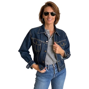 May include: A woman wearing a dark blue denim jacket over a grey long-sleeved shirt and blue jeans. She is wearing sunglasses and has her hair pulled back.