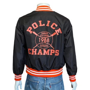 May include: Black satin baseball jacket with orange and white striped cuffs and waistband. The back of the jacket features a large graphic with the text "POLICE CHAMPS" and a baseball with crossed bats. The graphic also includes the text "HARRISON LITTLE LEAGUE 1988 EASTERN DIVISION".