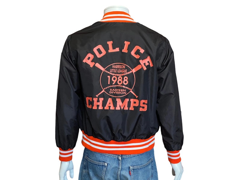 May include: Black satin baseball jacket with orange and white striped cuffs and waistband. The back of the jacket features a large graphic with the text "POLICE CHAMPS" and a baseball with crossed bats. The graphic also includes the text "HARRISON LITTLE LEAGUE 1988 EASTERN DIVISION".