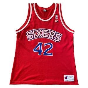 Puede incluir: Camiseta de baloncesto roja con ribetes blancos y azules. La camiseta tiene la palabra "SIXERS" en letras azules y blancas con un contorno azul. El número "42" está en azul con un contorno blanco. La camiseta tiene un parche del logotipo de Champion en la parte inferior derecha.