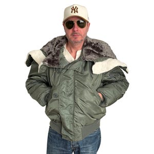 Può includere: Giacca bomber verde oliva con cappuccio foderato in pelliccia e orlo e polsini a coste. La giacca ha una chiusura con bottoni e due tasche anteriori. La persona indossa un berretto da baseball bianco con logo marrone e occhiali da sole da aviatore.