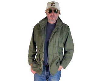 May include: An olive green field jacket featuring a high collar, four front flap pockets, and a front zipper closure. The jacket is worn over a dark blue shirt and paired with blue denim jeans. A cream-colored baseball cap with a brown NY logo is worn.