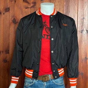 May include: Black satin baseball jacket with orange and white striped ribbing at the collar, cuffs, and waistband. The jacket has a button closure and features the word "Pam" embroidered on the left chest in orange thread.