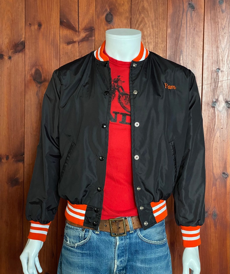 May include: Black satin baseball jacket with orange and white striped ribbing at the collar, cuffs, and waistband. The jacket has a button closure and features the word "Pam" embroidered on the left chest in orange thread.