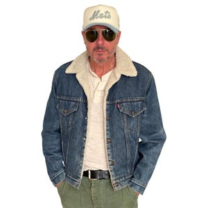 May include: A denim jacket with a cream-colored sherpa lining and a button-up front. The jacket is worn over a light-colored polo shirt and paired with olive-green pants. A baseball cap with the word "Mets" is worn.