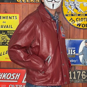 May include: A burgundy leather jacket with a button-up front, worn with blue jeans. The jacket has a collar and pockets. A black bowler hat and a white mask with a stylized face are also visible.