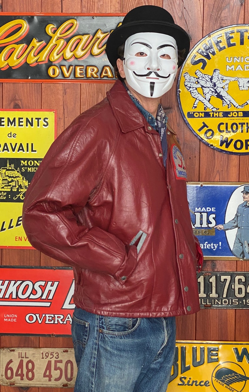 May include: A burgundy leather jacket with a button-up front, worn with blue jeans. The jacket has a collar and pockets. A black bowler hat and a white mask with a stylized face are also visible.