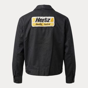 Könnte beinhalten: Schwarze Jacke mit Kragen und geknöpften Manschetten. Ein gelb-weißer Aufnäher auf dem Rücken zeigt "Hertz Quality Control."