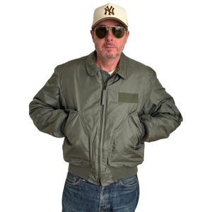 Può includere: Giacca bomber verde oliva con chiusura a zip, due tasche anteriori e una toppa sul petto. La giacca è abbinata a jeans blu e un berretto da baseball color crema con un logo. La persona indossa anche occhiali da sole da aviatore.