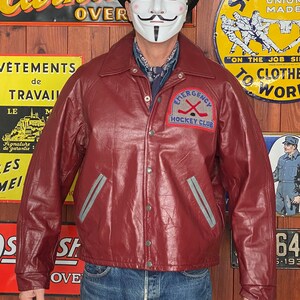 May include: Burgundy leather jacket with a gray stripe detail and a patch that reads "Emergency Hockey Club." The jacket has a button-up front and a collared neckline. The background includes vintage signs.