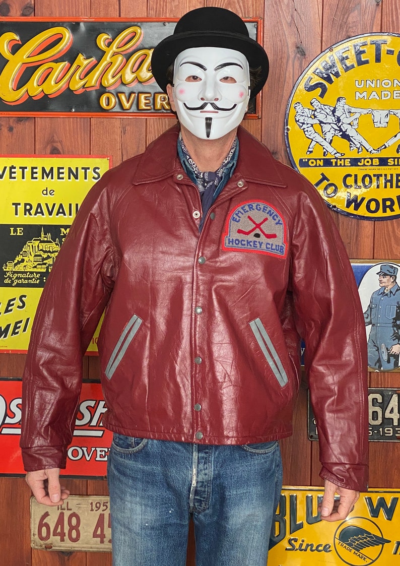 May include: Burgundy leather jacket with a gray stripe detail and a patch that reads "Emergency Hockey Club." The jacket has a button-up front and a collared neckline. The background includes vintage signs.