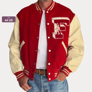 Op de afbeelding: Een rood varsity-jack met crèmekleurige mouwen, een geribbelde kraag en boorden met rode strepen. De voorkant toont een grote letter F met een kleine afbeelding en een label met maat 44 US. Het jack heeft een sluiting met drukknopen.