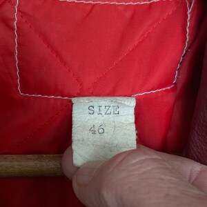 May include: Close-up of a red quilted fabric lining with a white stitched border. A tag with the word "SIZE" and the number "46" is visible. The tag is held by a person's hand.
