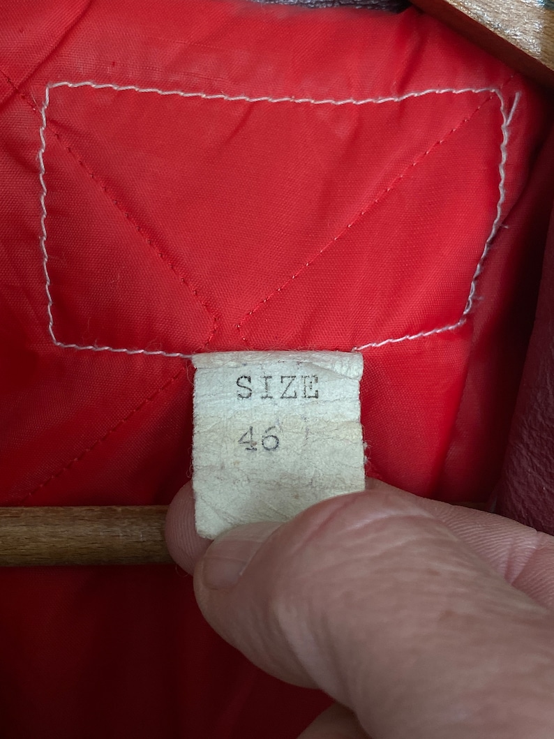 May include: Close-up of a red quilted fabric lining with a white stitched border. A tag with the word "SIZE" and the number "46" is visible. The tag is held by a person's hand.