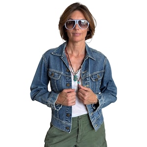 May include: A blue denim jacket with a button-up front, worn over a white top and green trousers. The outfit is accessorized with a necklace and light blue aviator sunglasses. The jacket is a classic style.