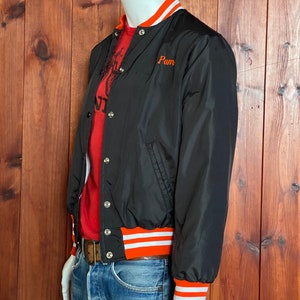 May include: A black bomber jacket with orange and white striped ribbing at the cuffs and hem. The jacket has a snap closure and a small embroidered patch that says "Pam".