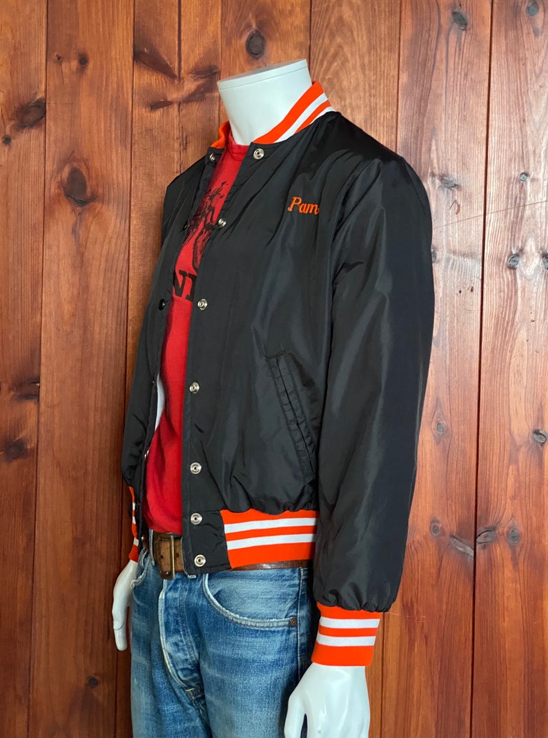 May include: A black bomber jacket with orange and white striped ribbing at the cuffs and hem. The jacket has a snap closure and a small embroidered patch that says "Pam".