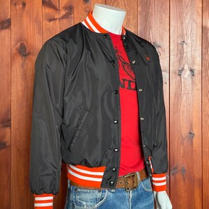 May include: A black satin bomber jacket with orange and white striped ribbing at the collar, cuffs, and waistband. The jacket has a snap closure and two front pockets.