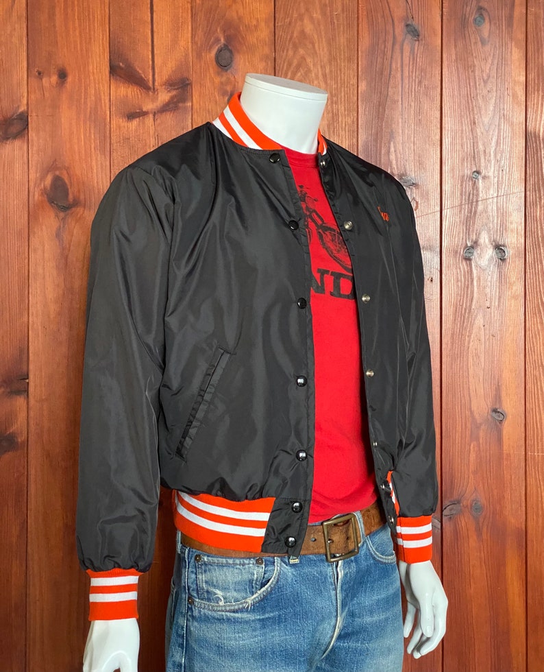 May include: A black satin bomber jacket with orange and white striped ribbing at the collar, cuffs, and waistband. The jacket has a snap closure and two front pockets.