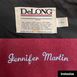 Op de afbeelding: Een close-up van een bordeauxrode jas met de naam "Jennifer Martin" geborduurd in lichtblauw garen. De binnenkant van de jas heeft een label met het merk "DeLONG" en wasvoorschriften. Het label vermeldt ook "Made in U.S.A."