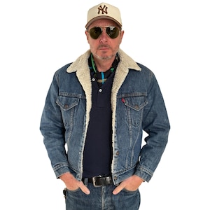 May include: A denim jacket with a cream-colored shearling collar and lining. The jacket is worn over a navy blue polo shirt and jeans. The person is wearing a baseball cap and sunglasses.