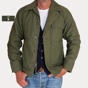 Puede incluir: Una chaqueta de color verde oliva con cuello en punta, cierre frontal de botones y un bolsillo en el pecho. La chaqueta tiene una etiqueta de talla S. Se lleva sobre una camiseta blanca, un chaleco azul marino a rayas y vaqueros azul claro.