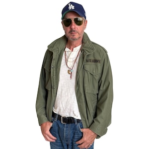 May include: A navy blue baseball cap with white LA logo, aviator sunglasses, an olive green field jacket with U.S. ARMY text, a white henley shirt, a multi-strand beaded necklace, and blue denim jeans with a black leather belt.
