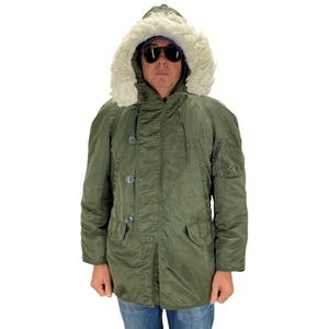 Può includere: Parka verde oliva con cappuccio foderato in pelliccia. Il cappotto invernale presenta un'abbottonatura frontale, tasche multiple e una vestibilità ampia. Il cappuccio ha un bordo in pelliccia color crema. La persona nell'immagine indossa occhiali da sole e jeans blu.