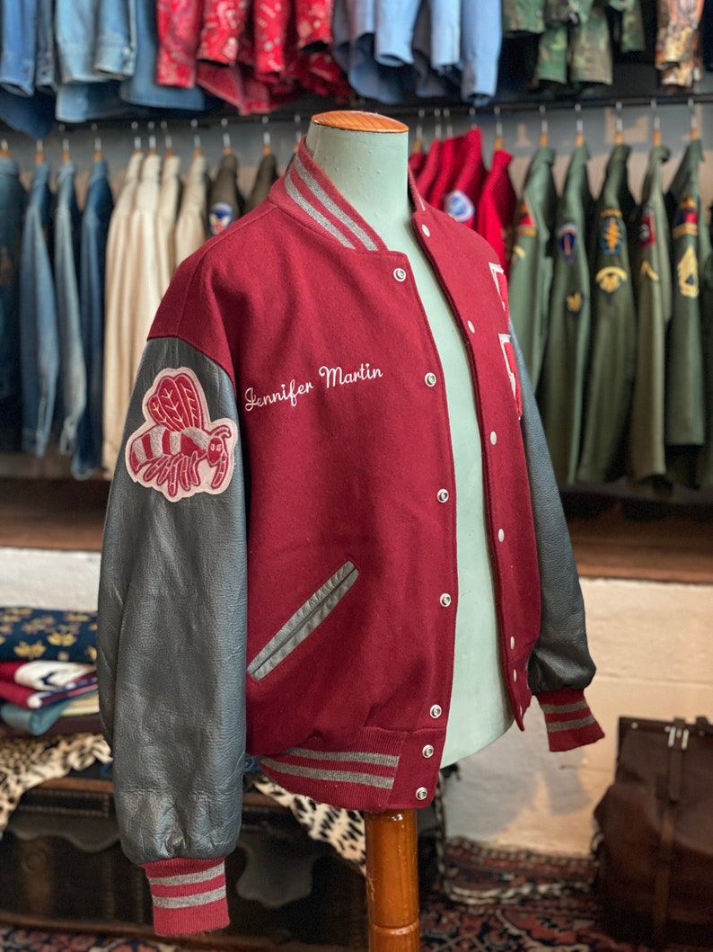Op de afbeelding: Bordeauxrode en grijze varsity-jas met zwarte mouwen. De jas heeft een bijenpatch op de linkermouw en de naam "Jennifer Martin" in het wit geborduurd. De jas heeft grijs-witte gestreepte manchetten en kraag.