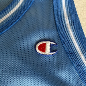 May include: A light blue sleeveless athletic jersey made of a breathable mesh fabric. The garment features a ribbed collar and armholes with white and blue striped trim. An embroidered Champion logo in red, white, and blue is on the chest.