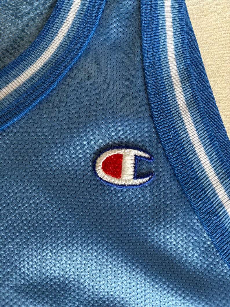 May include: A light blue sleeveless athletic jersey made of a breathable mesh fabric. The garment features a ribbed collar and armholes with white and blue striped trim. An embroidered Champion logo in red, white, and blue is on the chest.