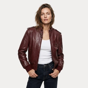 May include: A burgundy leather jacket with a ribbed hem and cuffs. The jacket has a zip closure and a small pocket on the chest. The model is wearing a white tank top and dark blue jeans. The jacket has a classic bomber style.