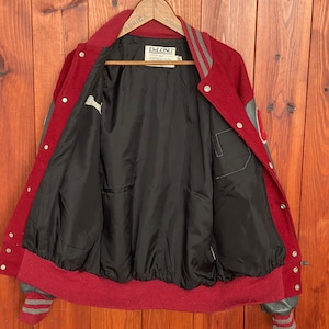 Op de afbeelding: Een vintage bordeauxrode en grijze varsity-jas met een zwarte voering. De jas heeft grijze mouwen, een bordeauxrode kraag en manchetten en een drukknoopsluiting. Het binnenlabel luidt "DeLONG".