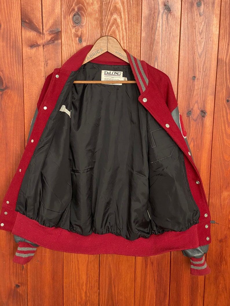 Op de afbeelding: Een vintage bordeauxrode en grijze varsity-jas met een zwarte voering. De jas heeft grijze mouwen, een bordeauxrode kraag en manchetten en een drukknoopsluiting. Het binnenlabel luidt "DeLONG".