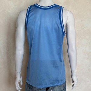 May include: A light blue sleeveless basketball jersey made of a breathable mesh fabric. The garment features a round neckline and armholes trimmed with white and dark blue stripes. The jersey is shown from the back on a mannequin torso.