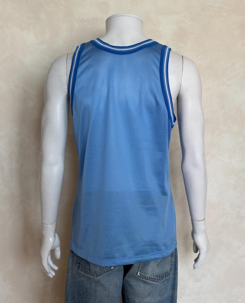 May include: A light blue sleeveless basketball jersey made of a breathable mesh fabric. The garment features a round neckline and armholes trimmed with white and dark blue stripes. The jersey is shown from the back on a mannequin torso.