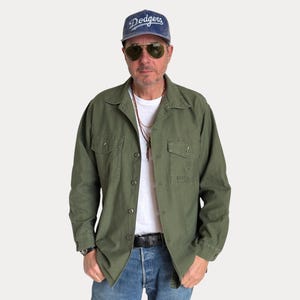 May include: An olive green button-down utility shirt with two chest pockets and USMC lettering, worn over a plain white t-shirt. The outfit includes blue denim jeans with a black belt, a blue Dodgers baseball cap, and gold-rimmed aviator sunglasses.