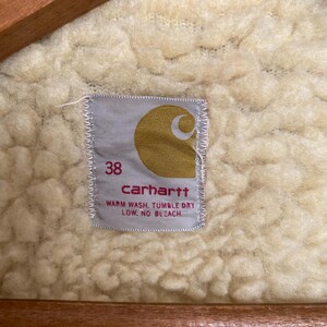 May include: A white Carhartt label with a brown logo and the text "carhartt" in black. The label also includes the size "38" and the care instructions "Warm wash, tumble dry low, no bleach."