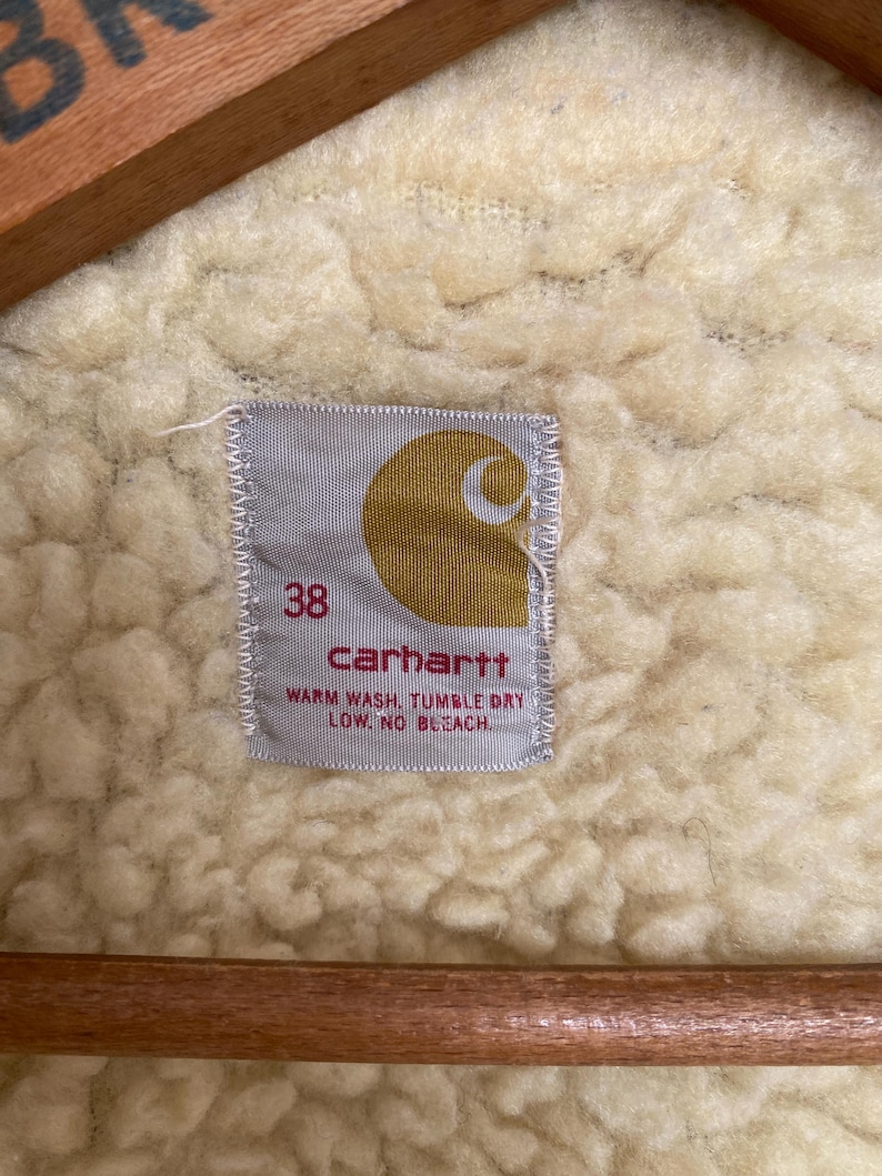 May include: A white Carhartt label with a brown logo and the text "carhartt" in black. The label also includes the size "38" and the care instructions "Warm wash, tumble dry low, no bleach."