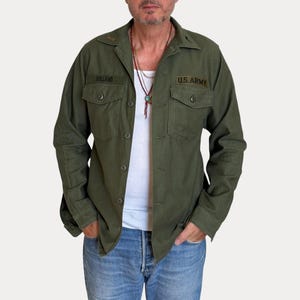 Puede incluir: Una camisa de manga larga abotonada en color verde oliva con dos bolsillos en el pecho con solapas. La camisa muestra una etiqueta con el nombre HOLLAND bordada en negro a la derecha y un parche negro U.S. ARMY sobre el bolsillo izquierdo.