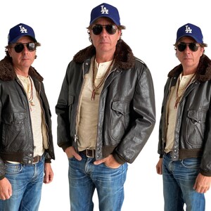 Puede incluir: Tres hombres con chaquetas de cuero marrón con cuellos de piel y gorras de béisbol azules con el logotipo de LA. Todos llevan pantalones vaqueros azules y camisetas blancas.